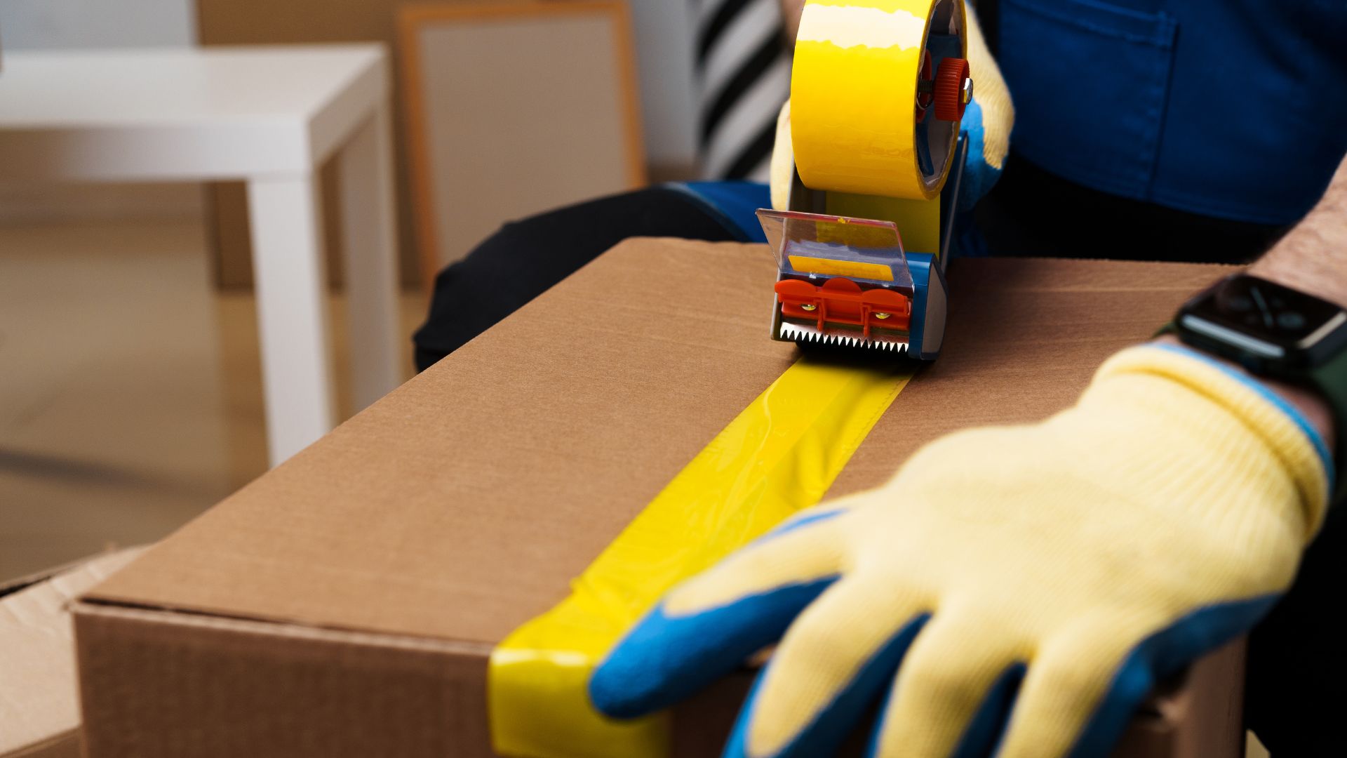 A person wearing yellow gloves and blue gloves is holding a box with a yellow tape